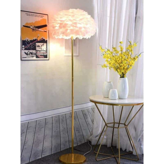 Lampadarul cu abajur din pene albe și tijă aurie, lângă o măsuță rotundă cu vaze și flori galbene